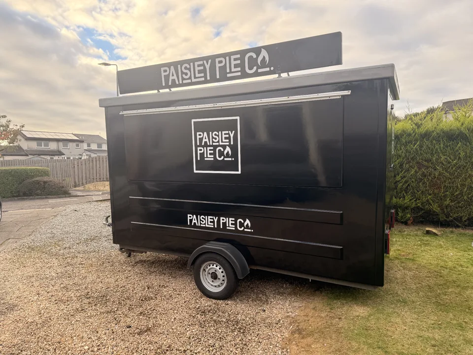 Completed black Paisley Pie Co trailer with white logos on the serving hatch, roof sign and lower side panel.